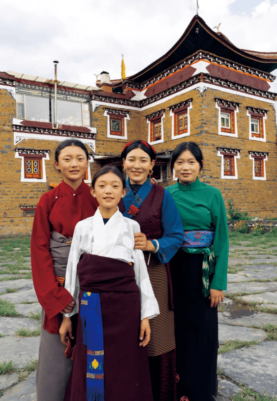 Four Tibetan women in traditional chuba dresses posing in front of an ornate stone building.