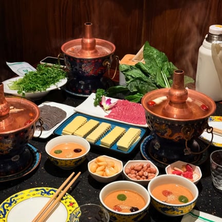 Traditional Chinese hot pot feast with copper pots, raw sliced beef, greens, and dipping sauces on a table.