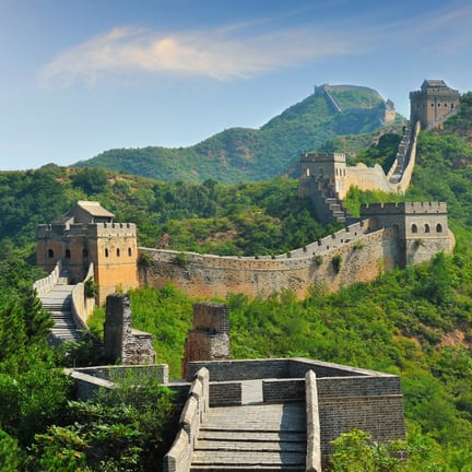 Ancient Great Wall of China winding through lush green mountains under a bright blue sky.