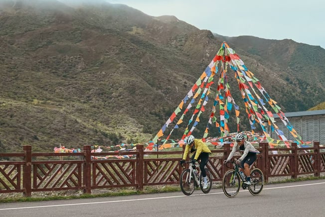 Two cyclists riding road bikes on a mountain pass decorated with colorful Tibetan prayer flags.