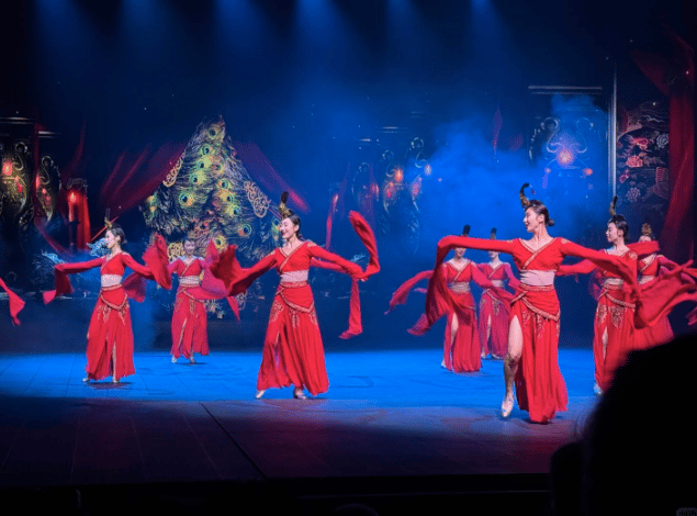 Dancers in vibrant red traditional Chinese costumes perform a folk dance on a stage with blue lighting.