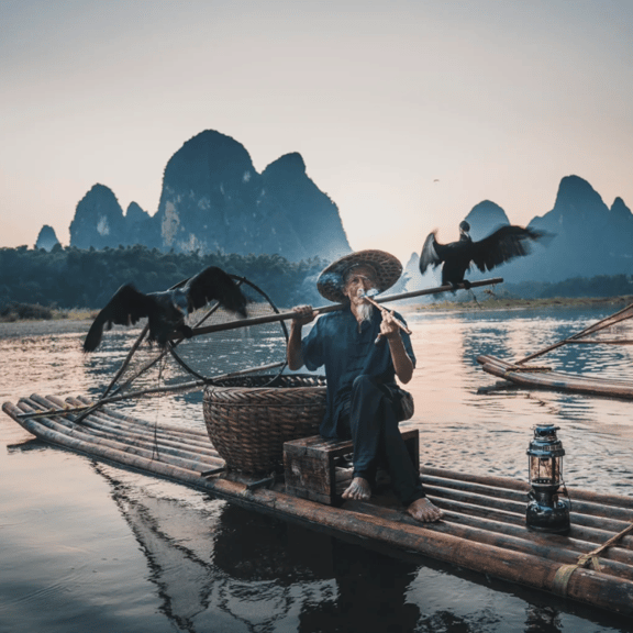 A traditional cormorant fisherman smoking a pipe on a bamboo raft at sunset in Yangshuo, China.