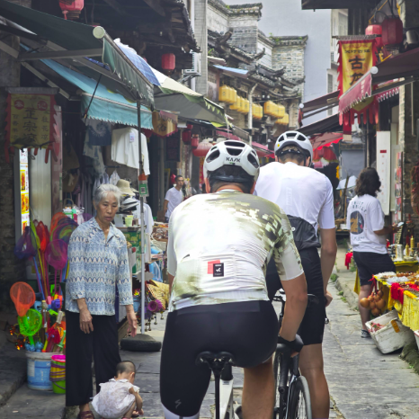 Two cyclists in professional gear ride through a narrow, traditional Chinese market street with local vendors and lanterns.