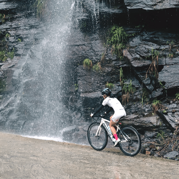 A female cyclist on a white road bike riding past a scenic waterfall on a rocky mountain road.
