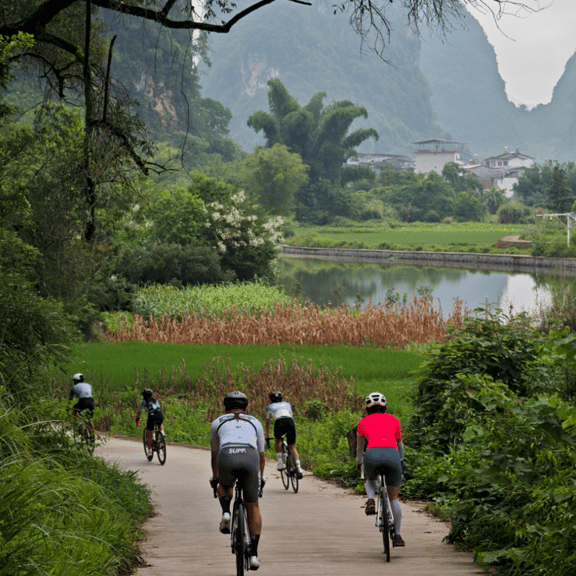 Cyclists riding road bikes on a paved path through a lush green valley with limestone mountains.