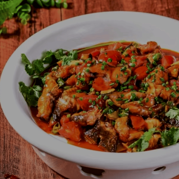 A white bowl of spicy Chinese braised fish with tomatoes, chives, and cilantro on a wooden table.