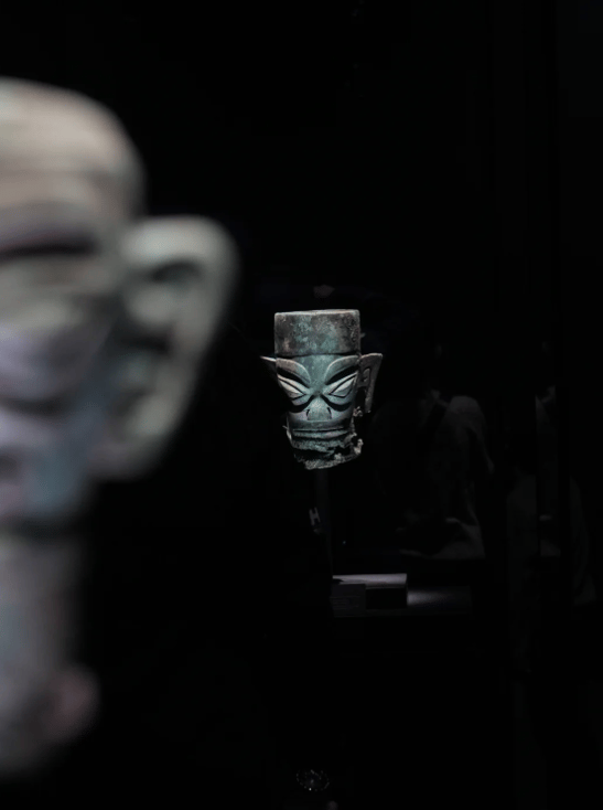 Ancient Chinese Sanxingdui bronze mask artifact displayed on a dark background in a museum.