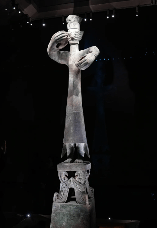 Ancient Sanxingdui bronze standing figure with large hands and a crown on display in a museum.