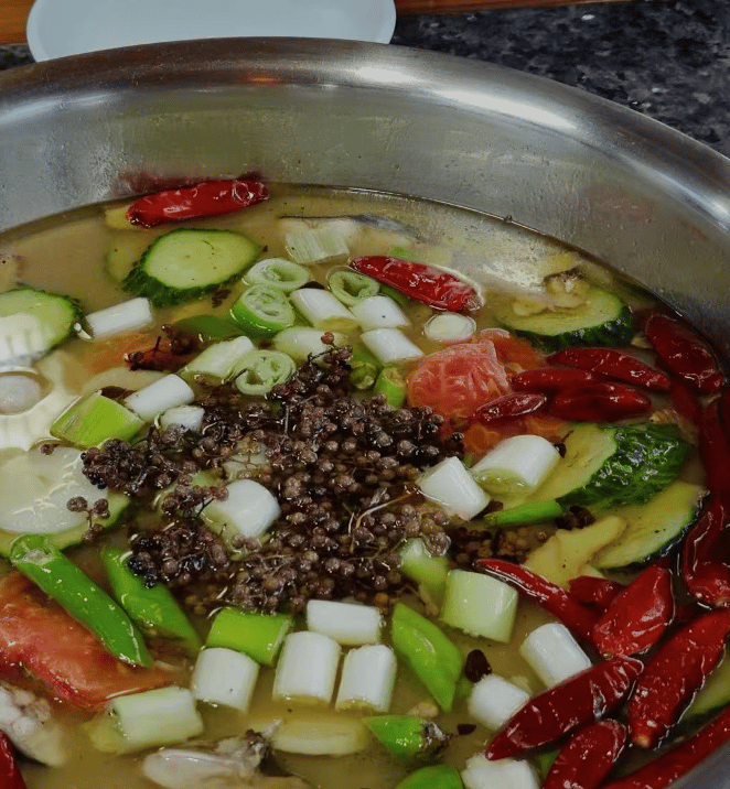 Spicy Chinese fish soup with Sichuan peppercorns, dried red chilies, scallions, and sliced cucumbers in a metal pot.
