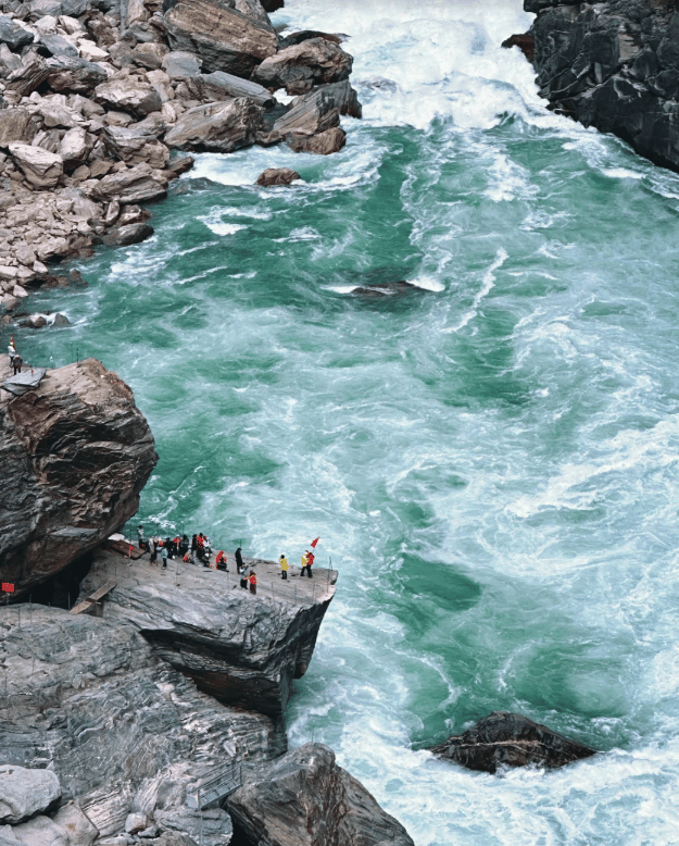 Tourists standing on a rocky viewing platform overlooking the rushing turquoise waters of Tiger Leaping Gorge.