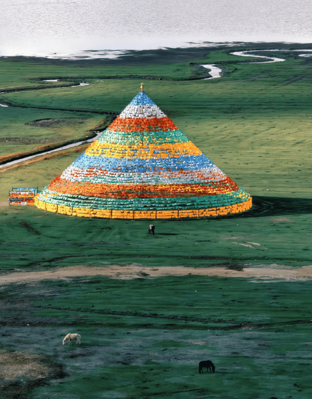 A conical structure draped in colorful Tibetan prayer flags stands on a green grassy plain near a river.